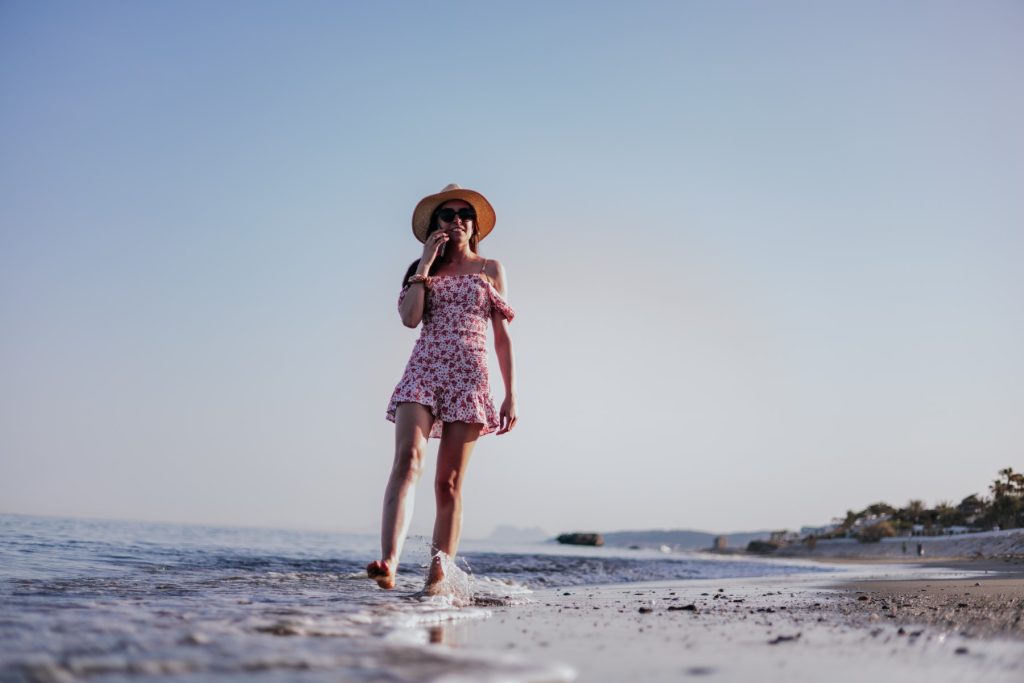 Brunette woman in red dress, hat and sunglasses talking happily with someone over phone, walking on beach